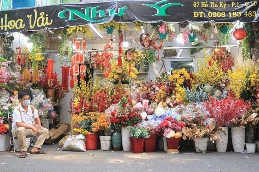 A flower shop with a variety of colorful flowers on display, a person sitting on a chair, and a sign with contact information. 4