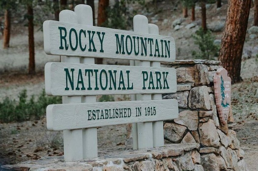 A wooden sign with the text 'ROCKY MOUNTAIN NATIONAL PARK ESTABLISHED IN 1915' in a natural setting. 1