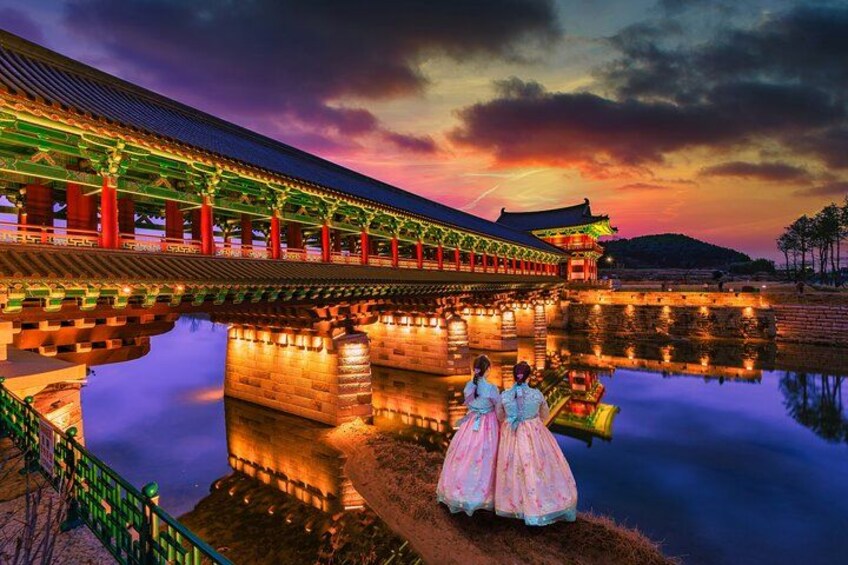 A traditional Korean palace with a reflection in the water, a bridge, and a person in a pink dress. 1