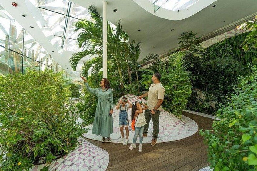 A family in a modern indoor garden with a wooden floor, surrounded by lush greenery and a glass ceiling. 3