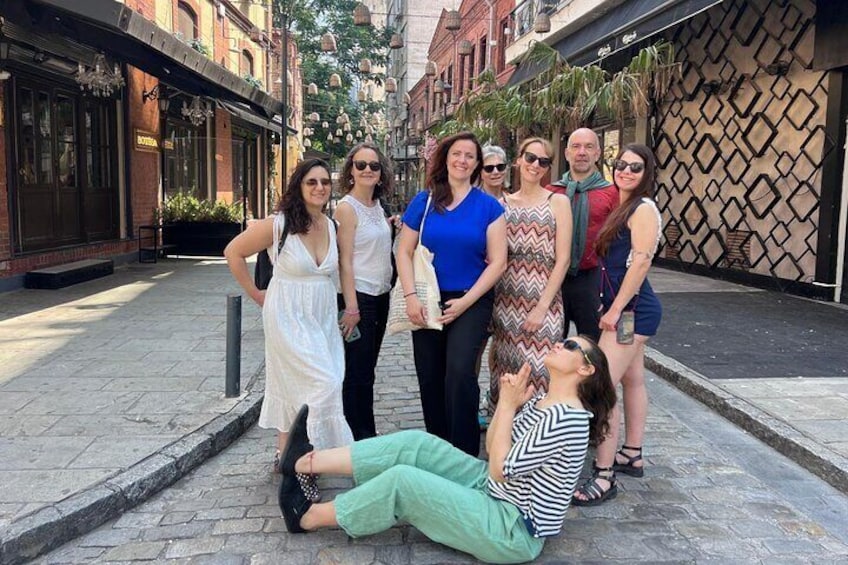 A group of people posing on a cobblestone street with buildings and a patterned wall in the background. 1
