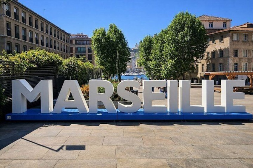 A large white sign with 'MARSEILLE' in capital letters on a blue base, set against a backdrop of buildings and trees. 2