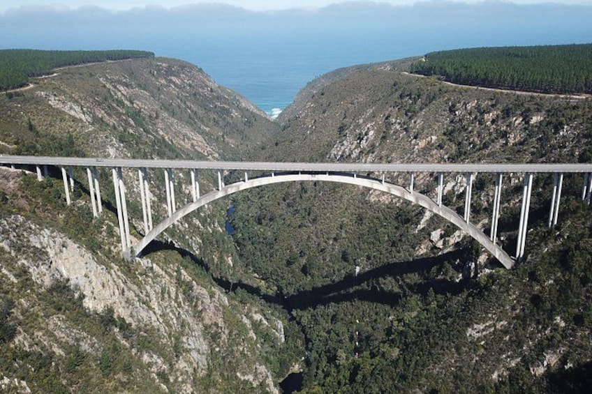 Eine Brücke überspannt ein tiefes Tal mit einer gebirgigen Kulisse und einem Gewässer in der Ferne. 1