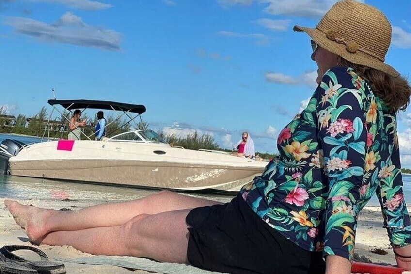 A person sitting on a dock by a boat, a sandy beach, and another boat in the background. 2