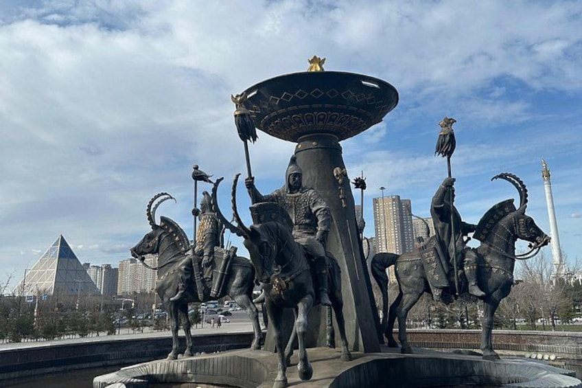 A large statue of a warrior on horseback with a shield, a sword, and a bird perched on the shield. In the background, there is a tall white monument and a pyramid-shaped building. 5