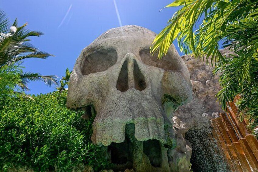A large stone skull sculpture surrounded by greenery and a clear blue sky. 3