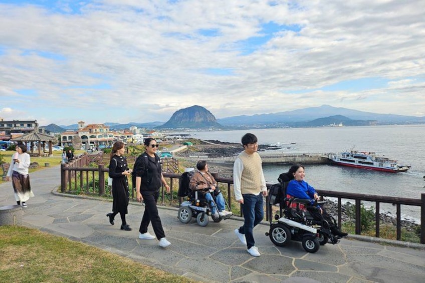 Un paseo marítimo con gente caminando, una silla de ruedas y un barco al fondo. 4