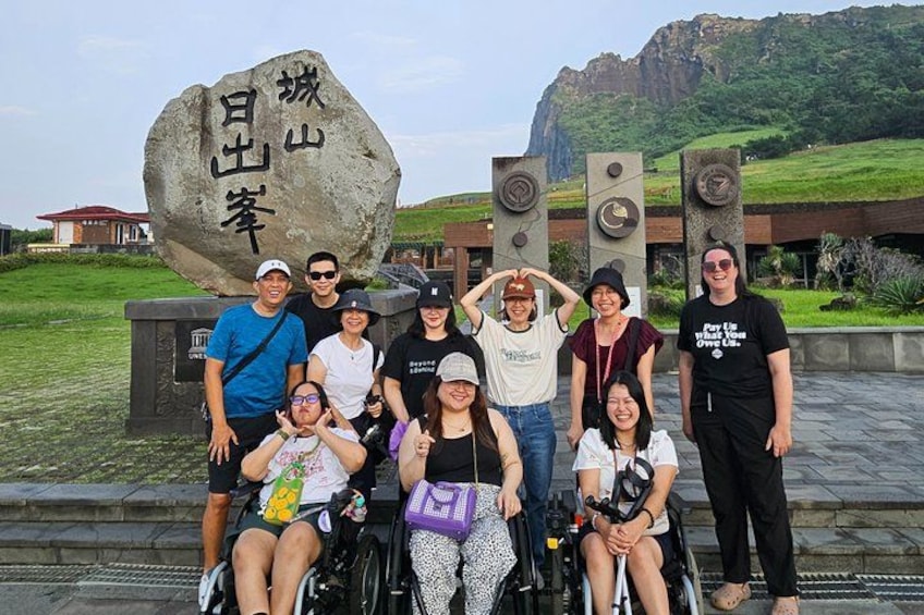 Un grupo de personas posando frente a una gran roca con inscripciones, un edificio moderno y un paisaje montañoso al fondo. 2