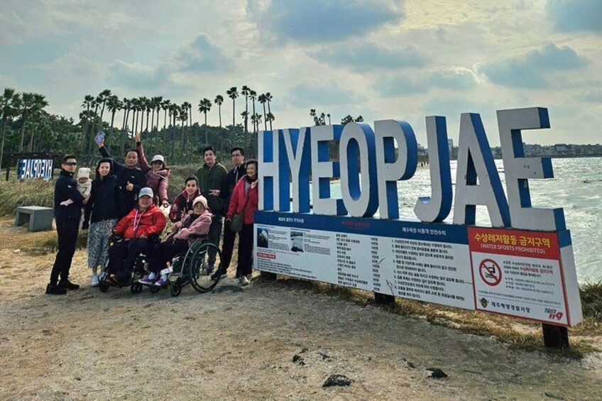 Un grupo de personas posando frente a un cartel que dice 'HYEOPJAE', con palmeras y una masa de agua al fondo. 3
