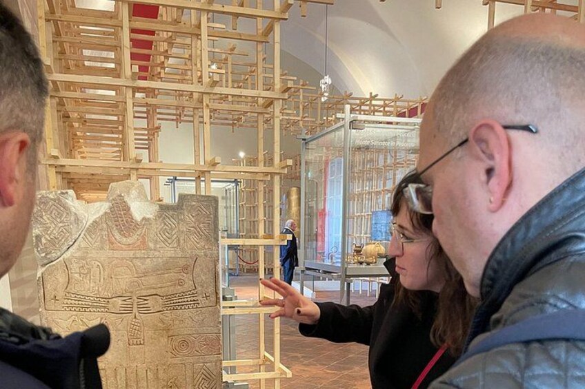 Et museumsinteriør med en stor, detaljeret udstilling, en person der peger på den, og en moderne geometrisk struktur i baggrunden. 3