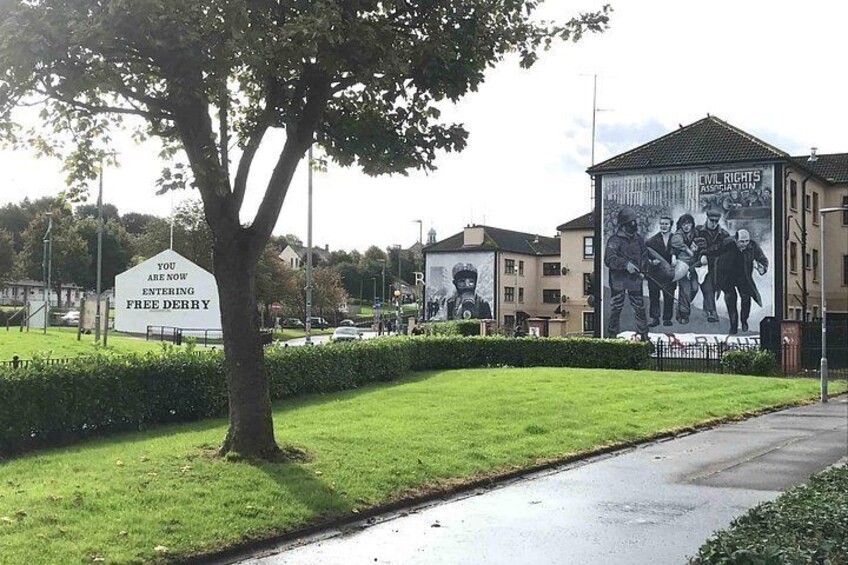 A tree-lined street with a sign indicating entry into Free Derry, a mural on a building, and residential houses. 2