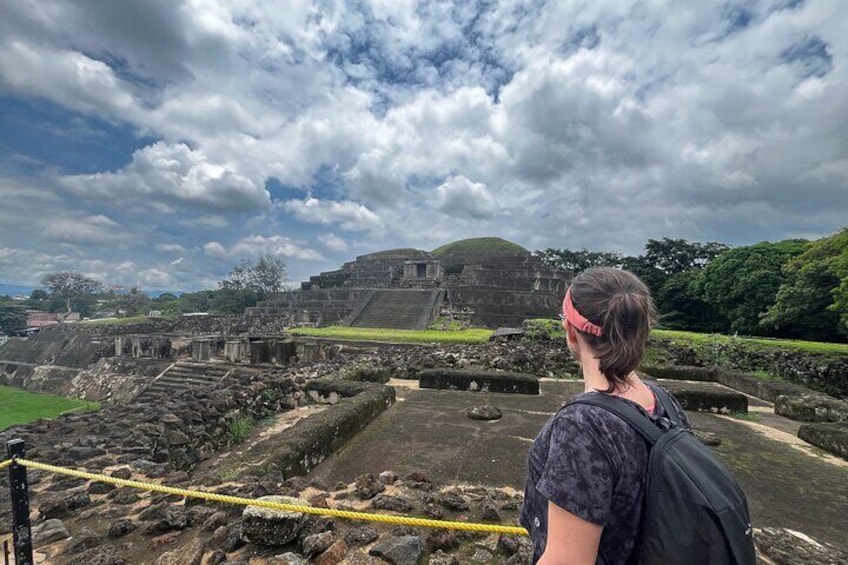 A person standing at an archaeological site with ancient ruins and a large mound in the background. 1