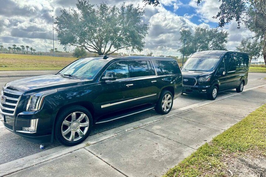 A black SUV and a black minivan parked on a concrete driveway with trees and a grassy field in the background. 4
