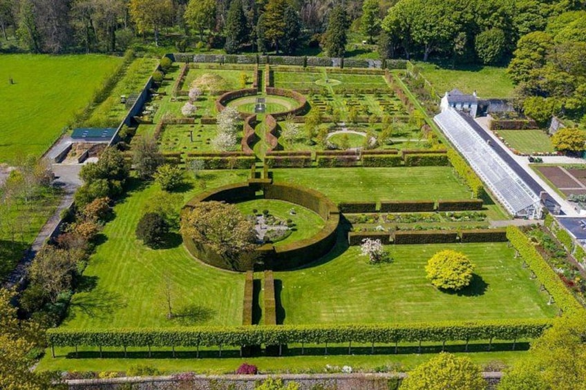 An aerial view of a formal garden with geometric patterns, a central circular feature, and a long building on the right. 1