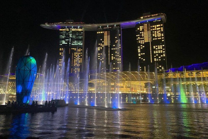A nighttime cityscape with illuminated buildings and a water fountain display. 1