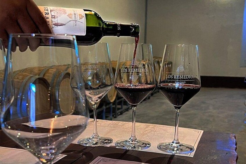 A bottle of wine being poured into three glasses labeled 'LOS REMEDIOS' on a table with a wooden background. 3
