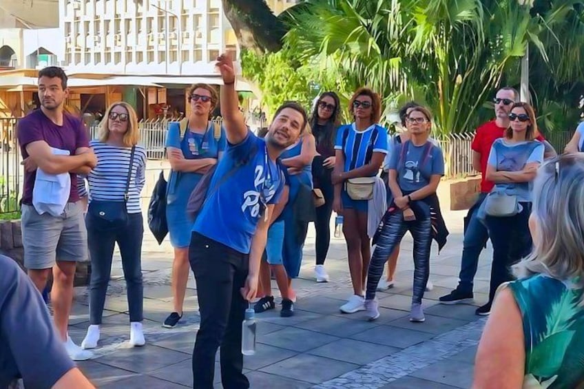 Un grupo de personas de pie al aire libre, algunas con los brazos cruzados y otras con las manos levantadas, en un área soleada y pavimentada con palmeras y edificios al fondo. 2