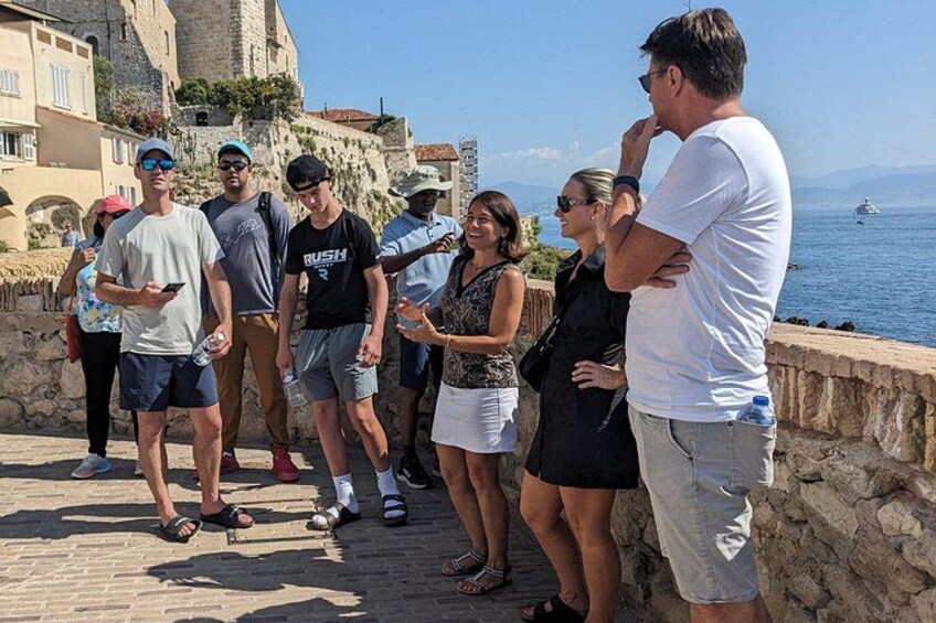 A group of people standing on a stone platform by the sea, with buildings and a tower in the background. 4