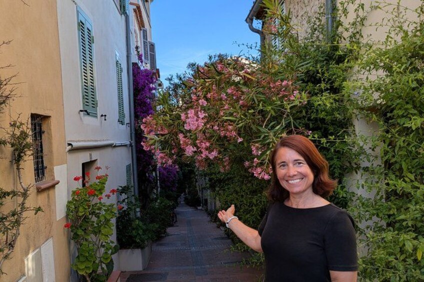 A narrow alley with buildings on either side, covered with flowering plants and vines. 1