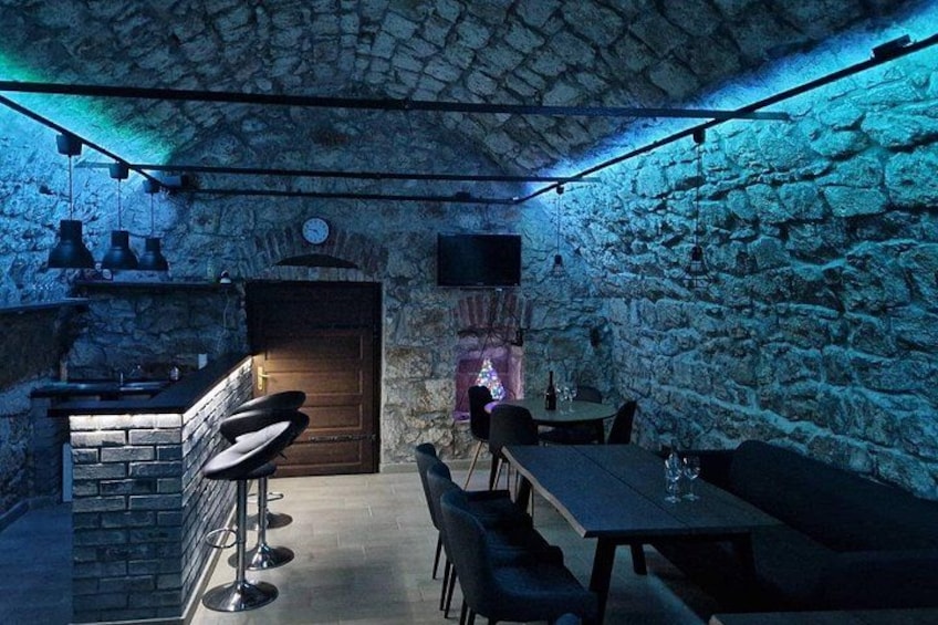 A rustic stone-walled room with a wooden door, bar stools, a table, and hanging lights. 5
