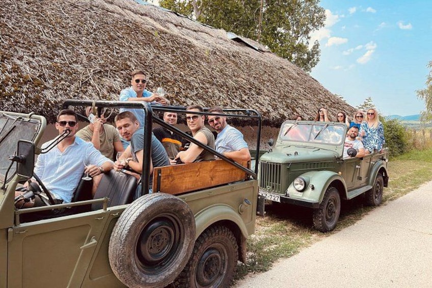A group of people in a vintage jeep on a rural road with a thatched-roof structure in the background. 1