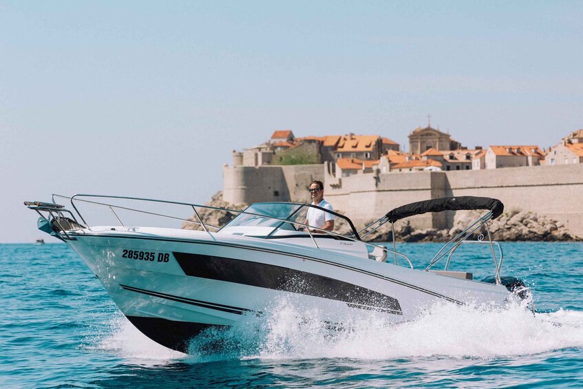 A speedboat with the number 285935 DB moving through clear blue water, with a coastal town and a large stone wall in the background. 3