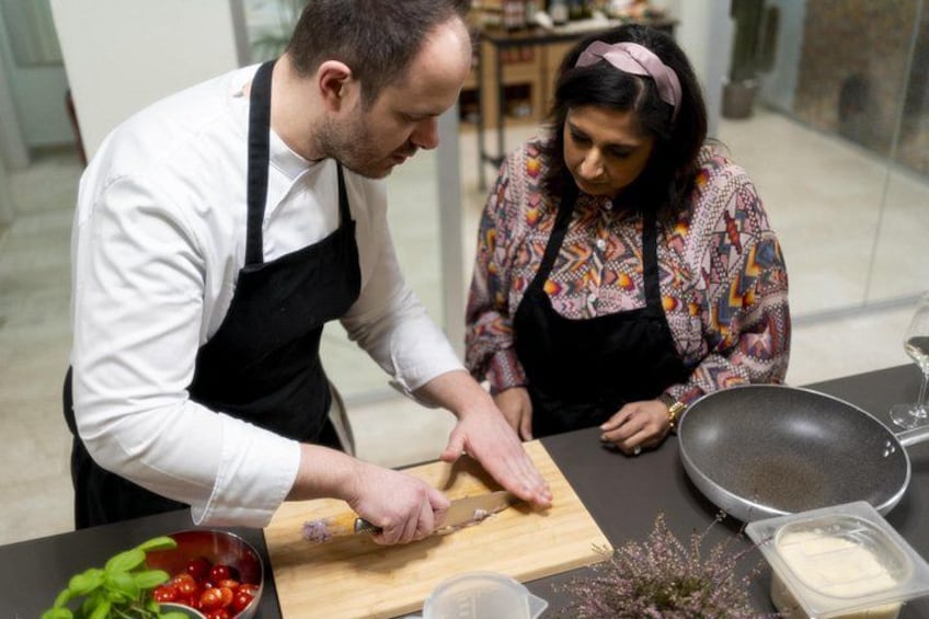 Two people preparing food, one cutting vegetables on a wooden board, with a bowl of tomatoes, a cutting board, and a frying pan on the table. 3