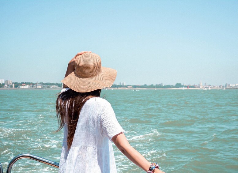 A person wearing a straw hat and a white dress, standing on a boat with a view of a body of water and a distant city skyline. 2
