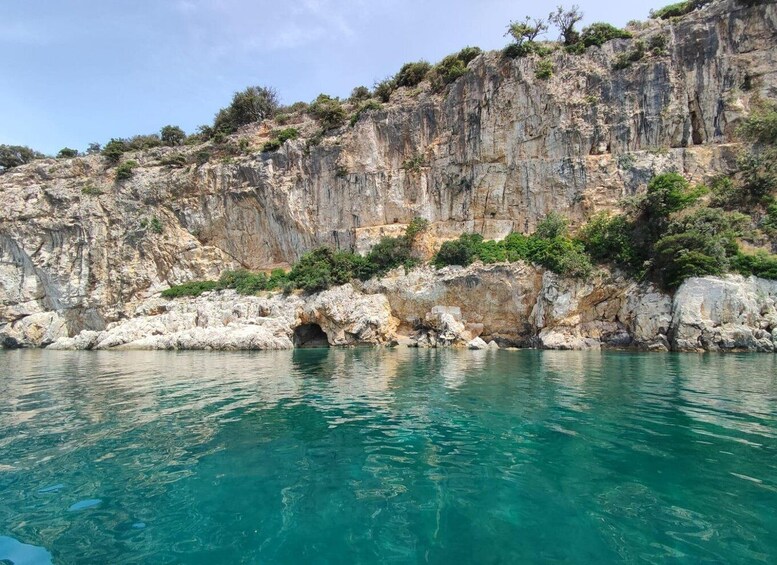 A coastal landscape with a rocky cliff, clear turquoise water, and a small cave entrance. 4