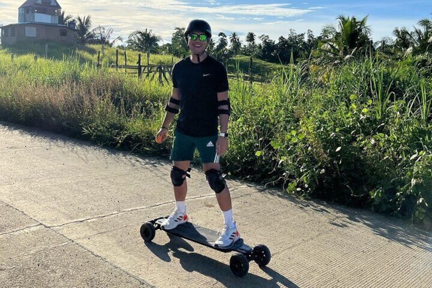 A person on a hoverboard on a paved path with greenery and a building in the background. 1