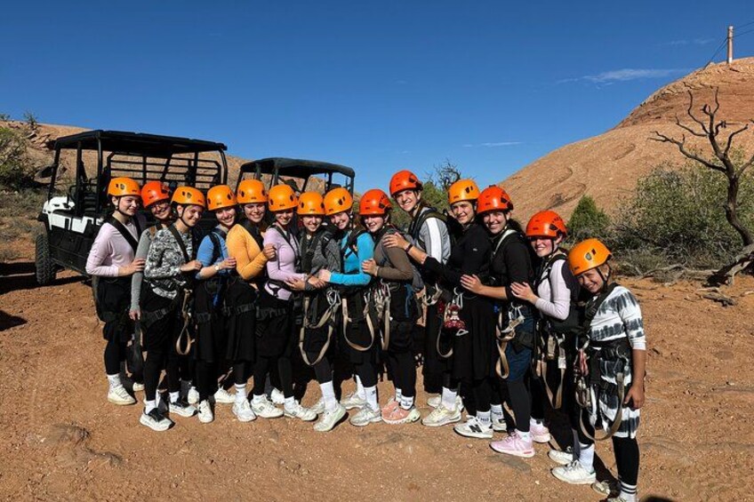 A group of people wearing helmets and harnesses, standing in a desert landscape with a vehicle and a hill in the background. 2