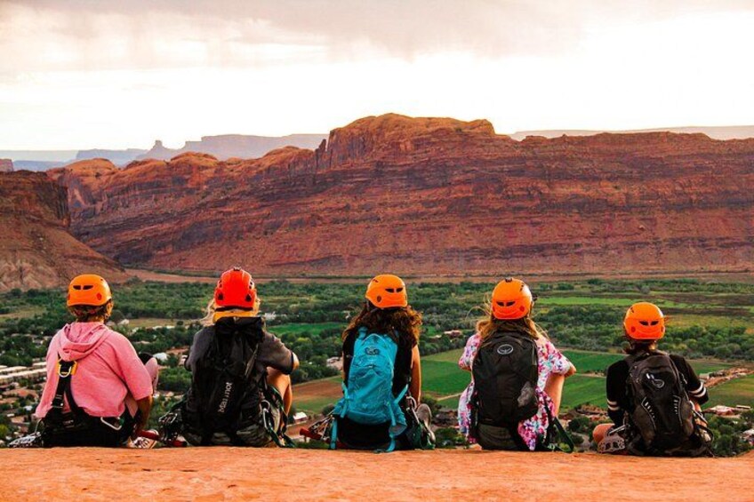 A group of people sitting on a ledge overlooking a canyon with red rock formations. 3