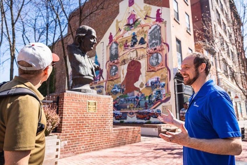 Un hombre con camisa azul conversando con un niño y un adulto cerca de una estatua y un mural. 3