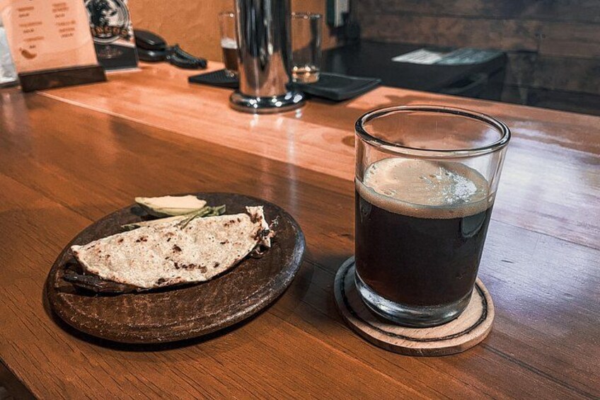 Een houten tafel met een glas donker bier en een plat, broodachtig gerecht op een onderzetter. 2