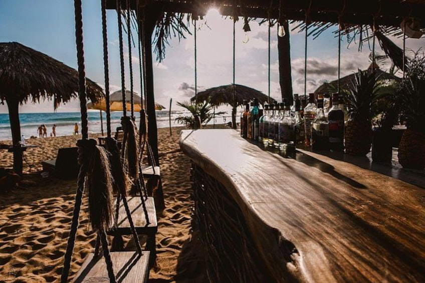 A beach bar with a wooden counter, thatched umbrellas, and a sandy beach in the background. 4