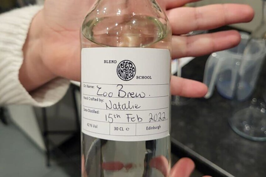 Een hand houdt een fles wijn vast, met een etiket waarop de naam 'Zoo Brew', de distilleerder 'Natalie' en de datum '15 februari 2022' staan. 3