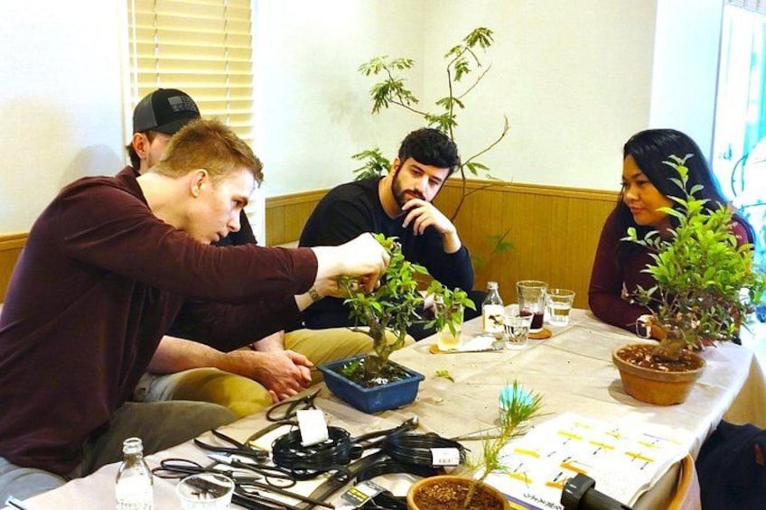 A group of people engaged in a plant-related activity at a table with potted plants, gardening tools, and papers. 3