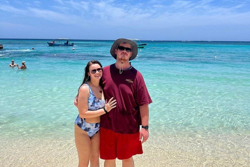 Et par står på en strand med krystalklart turkis vand og både i baggrunden. 5