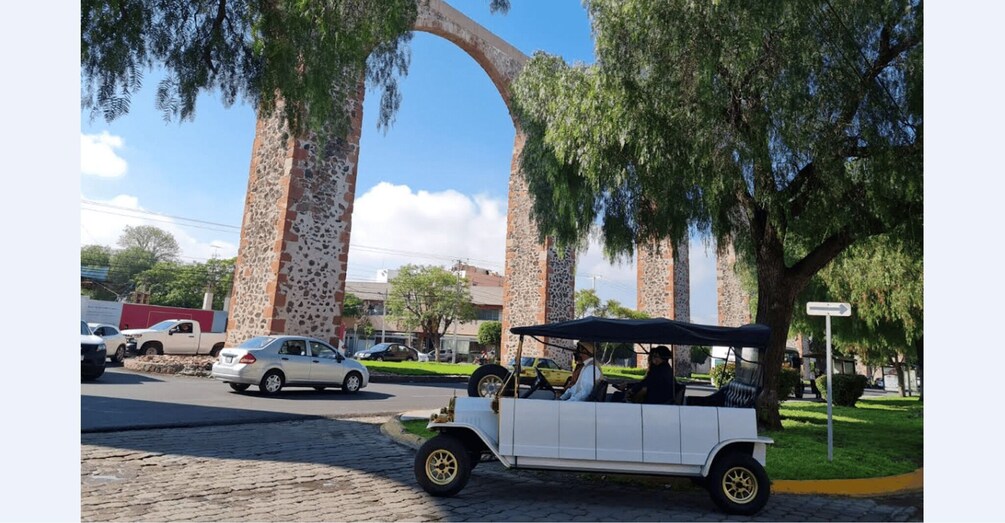 A white convertible car parked on a cobblestone street, a stone archway, and a golf cart in the background. 1