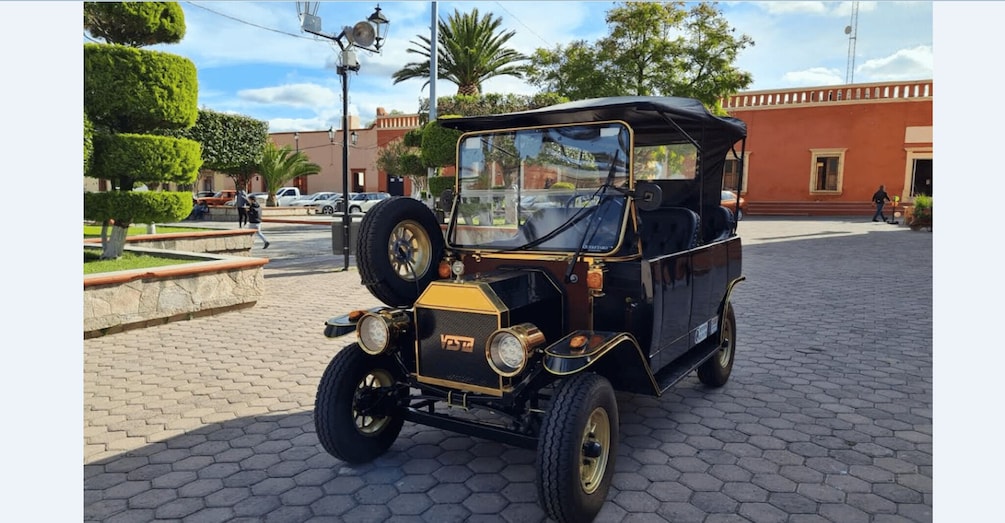 A vintage car parked on a cobblestone street with a building and trees in the background. 3