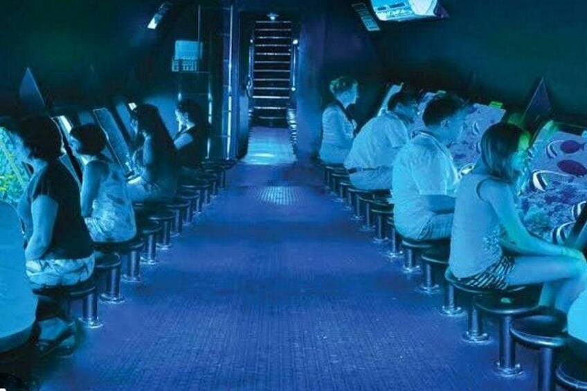 A row of people sitting on stools in a dimly lit room with a staircase in the background. 1