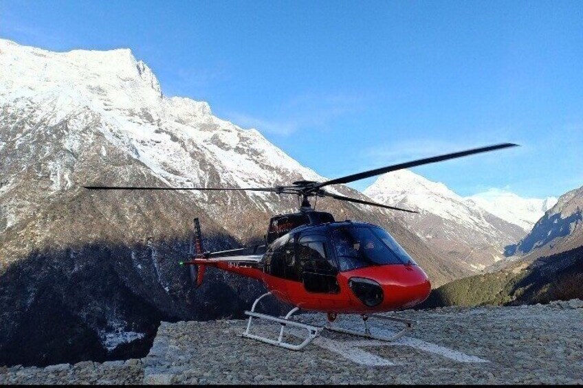 A red helicopter parked on a rocky surface with snow-capped mountains in the background. 1