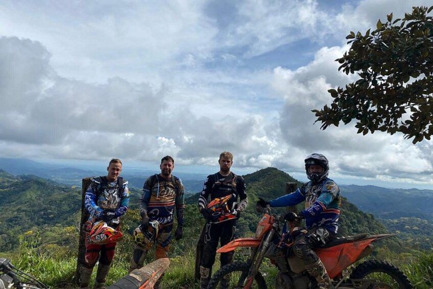 Quatre personnes sur une montagne, avec des motos, entourées de verdure et de nuages. 3