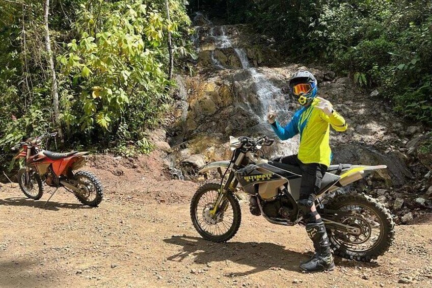 Une personne sur une moto tout-terrain dans une zone boisée, avec une cascade en arrière-plan. 4