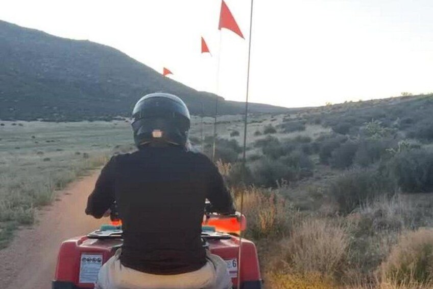 A person riding a quad bike on a dirt path with red flags in the distance and a hilly landscape. 3