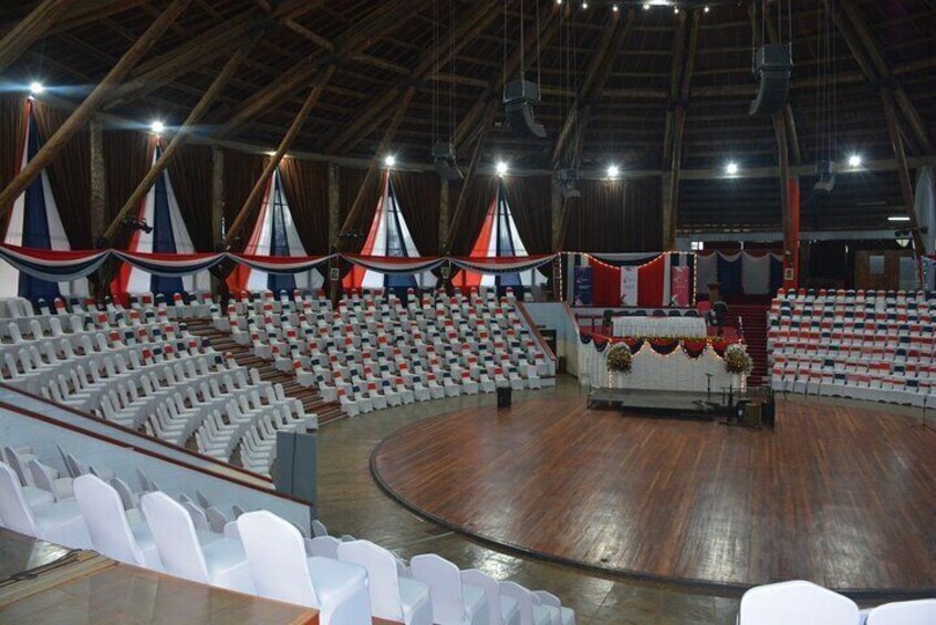 An indoor auditorium with tiered seating, a stage with a red, white, and blue backdrop, and a wooden floor. 3