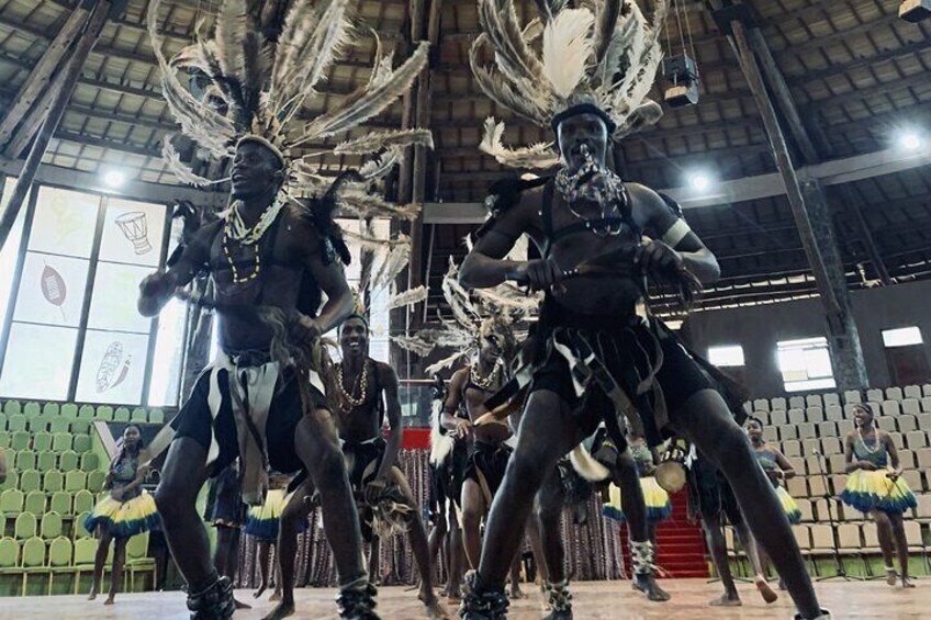 A group of dancers in traditional attire performing in an indoor venue with a high ceiling and green seating. 2