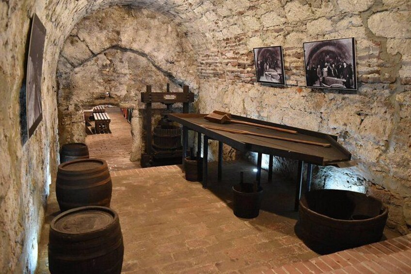 Une cave aux murs de pierre, avec des barils en bois, une longue table et deux tableaux encadrés accrochés au mur. 5