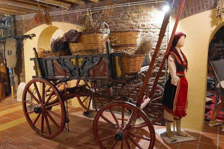 A vintage wooden carriage with red wheels, a mannequin dressed in traditional attire, and a rustic interior with stone walls and a tiled floor. 3