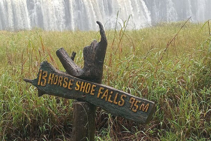 Un panneau indicateur en bois, sur lequel on peut lire « 13 Horse Shoe Falls 95 m », est placé devant une cascade et une zone herbeuse. 5
